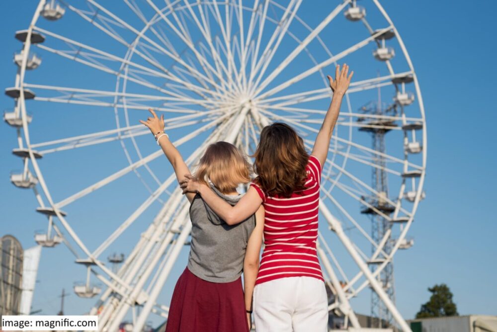 Mother and daughter with an arm around each other, and their other arm in the air, with their backs to the camera, facing a ferris wheel